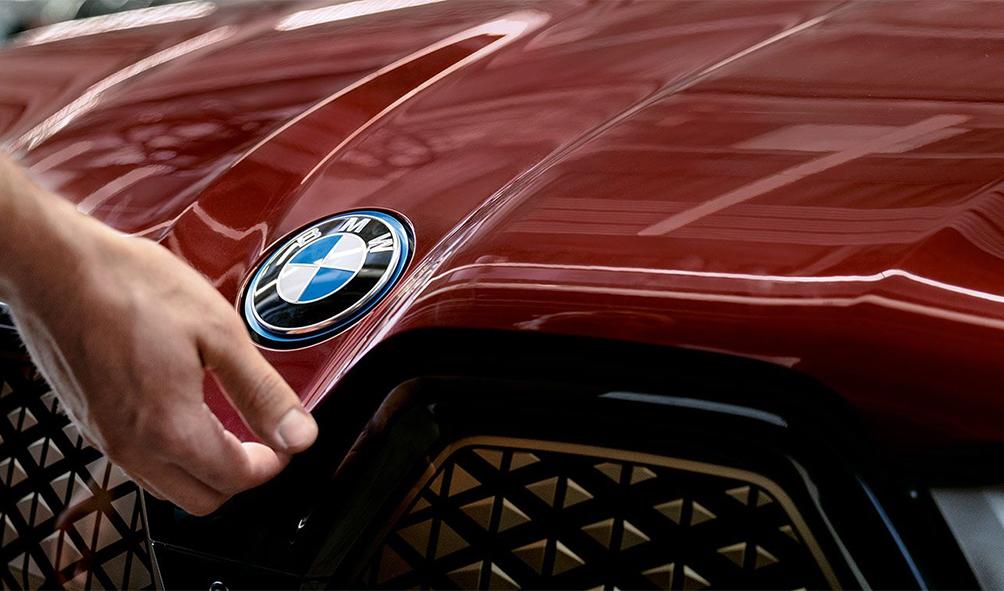 Das Bild zeigt eine Nahaufnahme der rotbraun glänzenden Motorhaube eines BMWs mit dem markanten BMW-Emblem in der Mitte, während eine Hand von links greift, um die Klappe unterhalb des Logos zu öffnen.