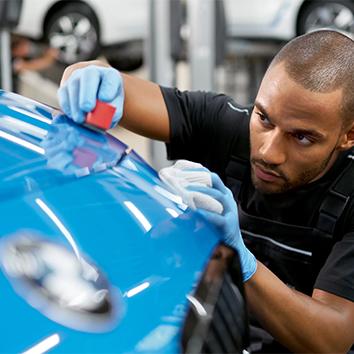 Ein Mechaniker mit blauen Schutzhandschuhen bessert sorgfältig mit Klotz und Tuch eine Lackstelle auf der Motorhaube eines blauen Autos aus.