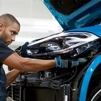 Ein Mechaniker mit Schutzhandschuhen montiert konzentriert den Scheinwerfer an der geöffneten Front eines blauen Autos.