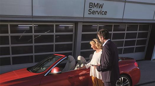 Frau und BMW Serviceberater stehen gemeinsam neben einem roten BMW Cabrio vor dem Servicebereich.