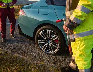 Pannenhelfer in Warnweste steht mit Werkzeug neben dem Hinterrad eines hellblauen BMW.