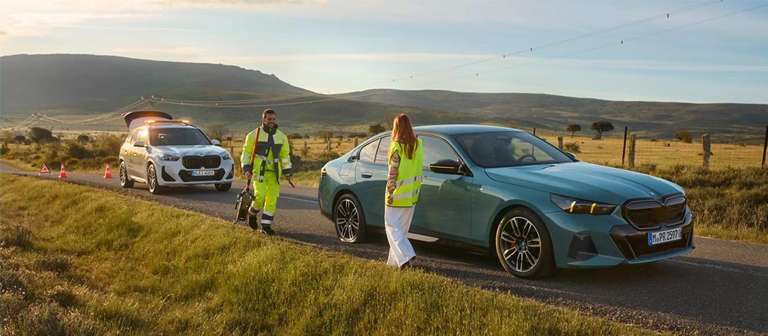 Pannenhelfer und Frau in Warnwesten stehen neben einem hellblauen BMW auf einer Landstraße, im Hintergrund ein BMW Servicefahrzeug.