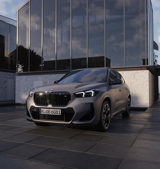 Das Bild zeigt einen sportlichen BMW X1 M35i in einer matten, grauen Lackierung. Das Kompakt-SUV steht auf einem modern gepflasterten Platz vor einem minimalistischen Gebäude mit einer großen Glasfassade, in der sich die Wolken spiegeln.