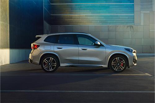 Der silberfarbene BMW X1 ist in einer Seitenansicht auf einem Parkplatz dargestellt. Im Hintergrund sind moderne Gebäudefassaden mit dunklen und hellblauen Akzenten zu sehen. Die Aufnahme betont die Seitenlinie und das Felgendesign des Wagens.