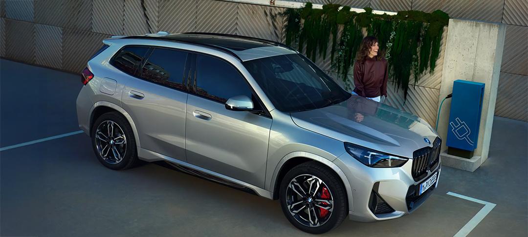 Ein silberfarbener BMW X1 steht auf einem Parkplatz neben einer blauen Ladesäule. Eine Frau lehnt am Auto und lächelt. Die Umgebung ist modern, mit einer strukturierten Wand, die mit Efeu oder anderen Pflanzen bewachsen ist. Die Aufnahme ist von schräg vorne-oben gemacht und präsentiert das Fahrzeug als Teil eines modernen, umweltbewussten Lebensstils.