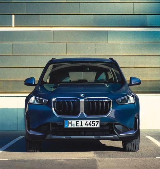 Das Bild zeigt die Frontansicht eines dunkelblauen BMW X1, vermutlich das Modell xDrive23i oder ähnlich, das in einer Parklücke steht.  Man sieht deutlich die typische, große, leicht eckige BMW-Doppelniere als Kühlergrill in der Mitte. Links und rechts davon befinden sich schmale, moderne Scheinwerfer. Das Auto wirkt muskulös und steht vor einer modernen Fassade, die aus horizontalen, graublauen Paneelen besteht. Das Licht ist eher weich. Auf dem Nummernschild steht 'M EI 4457'.