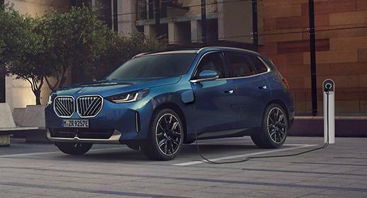 Das Bild zeigt den dunkelblauen BMW X3 von vorne links auf einem gepflasterten Parkplatz vor einem modernen Gebäude. Das Fahrzeug ist über ein schwarzes Kabel an einer minimalistischen, weißen Ladesäule angeschlossen, wobei die geöffnete Ladeklappe am vorderen linken Kotflügel gut sichtbar ist.