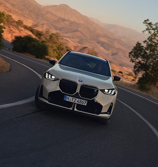 Das Fahrzeug präsentiert sich in einer dynamischen Kurvenfahrt vor einer malerischen Bergkulisse im warmen Abendlicht. Die Frontansicht wird durch die markante, beleuchtete Niere und die präzisen, vertikalen Lichtsignaturen der Scheinwerfer dominiert. In einem eleganten Beigeton gehalten, unterstreicht das SUV seine Rolle als sportliches Performance-Modell, das Souveränität mit Abenteuerlust verbindet.