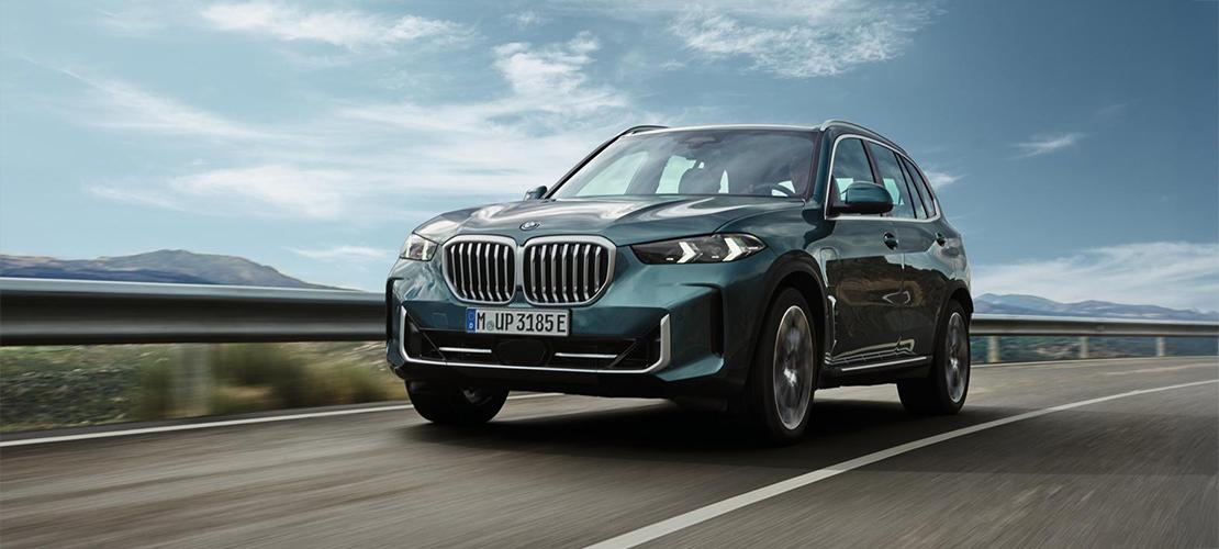 BMW X5 eDrive50 Aktionsangebot Ein Plug-in Hybrid BMW X5 in einer dunklen Petrol- oder Grünmetallic-Farbe fährt auf einer mehrspurigen Straße. Das Fahrzeug ist dynamisch von vorne-rechts aufgenommen, wodurch die markante BMW-Niere, die schmalen Scheinwerfer und die sportlichen Linien gut zur Geltung kommen. Die Landschaft im Hintergrund besteht aus verschwommenen Bergen und einem weiten Himmel mit einigen Wolken, was den Eindruck von Geschwindigkeit und Bewegung verstärkt. Das Kennzeichen des Fahrzeugs ist "M UP 3185 E".