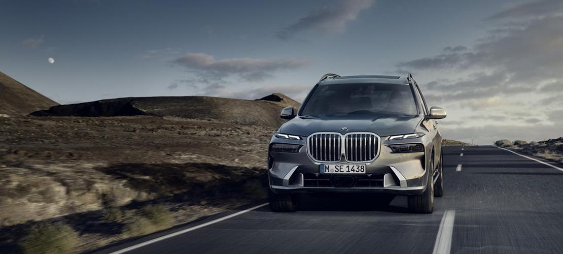 BMW X7 Angebot Ein dunkler, moderner BMW X7 SUV fährt frontal auf einer leeren Straße durch eine karge, bergige Landschaft. Der Himmel ist bewölkt, und ein heller Mond oder ein ähnliches Himmelsobjekt ist links oben sichtbar. Das Fahrzeug hat eine große, dominante Frontpartie mit dem charakteristischen BMW-Nierengrill und schlanken Scheinwerfern. Auf dem Kennzeichen steht "M SE 1438". Die Straße wirkt leer und erstreckt sich in die Ferne, was ein Gefühl von Weite und Erkundung vermittelt.