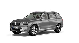 BMW X7 Das Bild zeigt einen grauen BMW X7. Es ist ein sehr großes, kastenförmiges SUV. Das Auto steht schräg von vorne auf einem weißen, leeren Hintergrund. Es hat einen großen vertikalen Kühlergrill und schlanke Scheinwerfer. Das Kennzeichen "M JE 9498" ist auf der vorderen Stoßstange zu sehen.