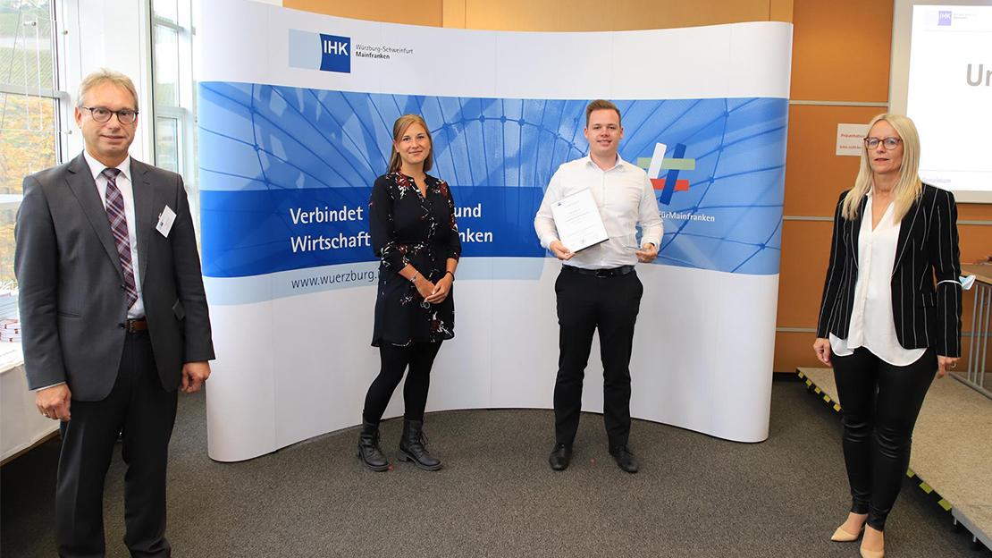 Vier Personen, darunter ein junger Mann mit einer Urkunde, stehen vor einer IHK-Wand.