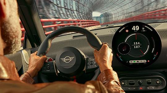 Bild präsentiert das innovative Herzstück des Interieurs: die MINI Interaction Unit, ein kreisförmiges OLED-Zentraldisplay mit 24 cm Durchmesser. In diesem modernen Cockpit verschmelzen Infotainment und Instrumentenanzeige zu einer Einheit, die durch das neue MINI Operating System 9 intuitiv per Touch oder Sprache gesteuert wird. Besonders markant ist das mit Textil bezogene Armaturenbrett, auf das Lichtprojektionen der verschiedenen „Experience Modes“ abgestimmt werden, um eine individuelle Atmosphäre zu schaffen. Die darunter liegende, reduzierte Schalterleiste zitiert das klassische MINI-Design und rundet das puristische, digitale Nutzererlebnis ab.