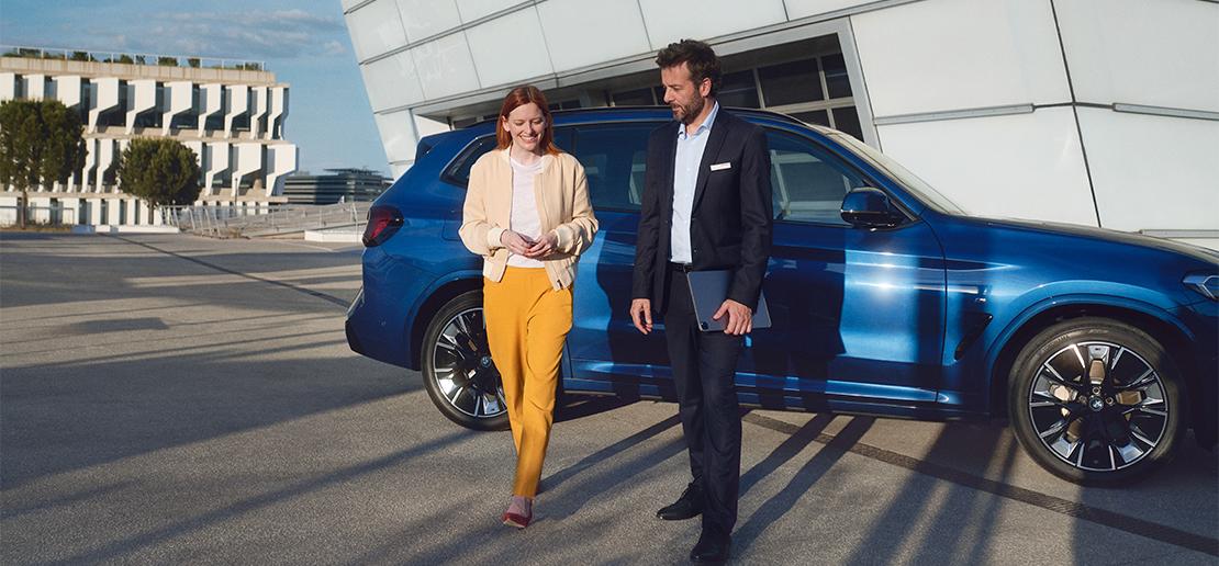 Fahrzeugankauf BMW Rhein Eine lächelnde Frau in gelber Hose und beige Jacke und ein Mann im Anzug mit Tablet gehen nebeneinander vor einem blauen BMW SUV und moderner Architektur.