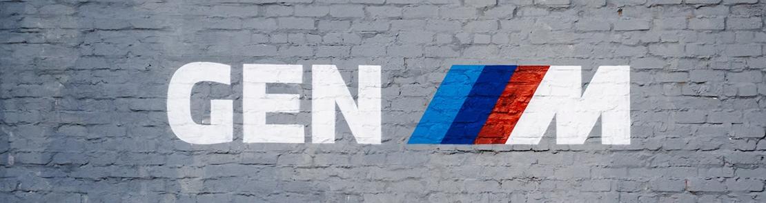 Das Wort "GEN M" ist in großen weißen Buchstaben auf einer grauen Ziegelwand gemalt. Zwischen "GEN" und "M" befinden sich drei diagonale Streifen in den Farben Hellblau, Dunkelblau und Rot, die das BMW M Logo repräsentieren.