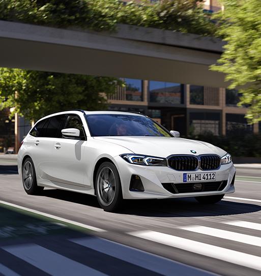 Das Bild zeigt einen weißen BMW 3er Touring (Kombi), der dynamisch über eine urbane Straße mit einem Zebrastreifen fährt. Der Wagen wirkt sportlich und modern, mit einer markanten Frontpartie, blauen Akzenten in den flachen LED-Scheinwerfern und einer klassischen, schwarzen Doppelniere. Die Umgebung ist durch grüne Bäume, eine moderne Betonbrücke im Hintergrund und helles, freundliches Tageslicht geprägt, was dem Bild eine lebendige, großstädtische Atmosphäre verleiht. Durch die leichte Bewegungsunschärfe an den Rädern und im Hintergrund wird ein Eindruck von Geschwindigkeit und Agilität vermittelt.