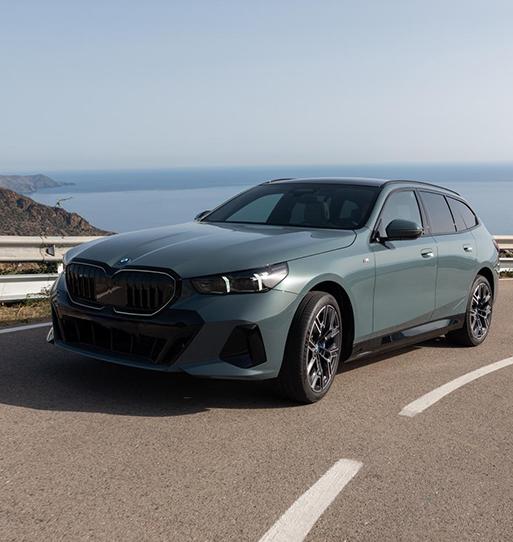 Das Bild zeigt einen modernen BMW 5er Touring in einem eleganten Grau-Blau mit metallischem Glanz. Der sportliche Kombi parkt auf einer sonnenbeschienenen Küstenstraße; im Hintergrund erstreckt sich ein ruhiges, hellblaues Meer unter einem klaren Himmel. Die Front des Wagens wird durch eine markante, schwarze Doppelniere und scharf geschnittene LED-Scheinwerfer geprägt, während große Alufelgen und bündige Türgriffe das aerodynamische Design betonen. Die gesamte Szenerie wirkt hochwertig und sommerlich, wie ein Moment während einer Urlaubsfahrt auf einer mediterranen Panorama-Route.
