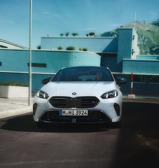 Dieses Bild präsentiert einen weißen BMW 1er in der sportlichen M-Ausführung aus einer direkten Frontperspektive. Das Fahrzeug wirkt durch die breite, schwarze Kühlermaske mit horizontalen Streben und die dunkel eingefassten Lufteinlässe besonders kraftvoll. Der Hintergrund ist architektonisch auffällig: Ein minimalistisches, hellblaues Gebäude mit klaren Linien und ein dahinter aufragender, karger Berg bilden einen starken Kontrast zum dynamisch fahrenden Auto auf der dunklen Asphaltstraße.