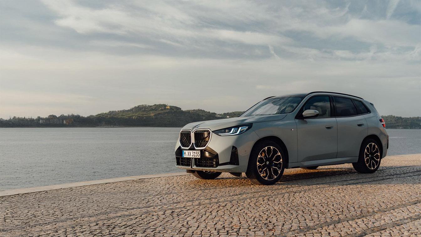 Das Bild zeigt ein hellgraues, modernes BMW SUV (vermutlich ein X1 oder X3) im Halbprofil von vorne rechts. Das Auto steht auf einem gepflasterten Uferweg direkt am Wasser, das sich nach links und in den Hintergrund erstreckt. Im Hintergrund ist eine bewaldete Hügellandschaft unter einem bewölkten Himmel zu sehen, der von der tief stehenden Sonne (wahrscheinlich zum Sonnenuntergang oder -aufgang) beleuchtet wird. Die Stimmung ist ruhig und leicht dramatisch. Das Fahrzeug ist sauber und poliert, mit schwarzen Felgen und eingeschaltetem Tagfahrlicht.