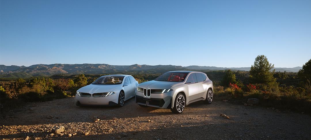 Das Bild zeigt zwei futuristische BMW-Konzeptfahrzeuge – links die Limousine BMW Vision Neue Klasse, rechts den SUV Vision Neue Klasse X – nebeneinander auf einem Schotterweg vor einer hügeligen Naturkulisse im warmen Abendlicht.