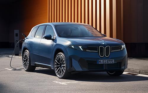 Dieses Bild zeigt den BMW iX3 in einer urbanen Ladesituation vor einer kontrastreichen Fassade aus vertikalen, orangefarbenen Lamellen. Die Frontansicht betont die beleuchtete, schmale Niere und die flachen Scheinwerfer, während das angeschlossene Ladekabel die Einsatzbereitschaft für den elektrischen Geschäftsalltag unterstreicht.