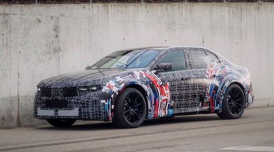 Eine getarnte BMW HP BEV M3 Limousine mit einem mehrfarbigen und schwarzen Tarnmuster steht auf einer Asphaltfläche vor einer grauen Betonwand. Das Fahrzeug hat schwarze Felgen.