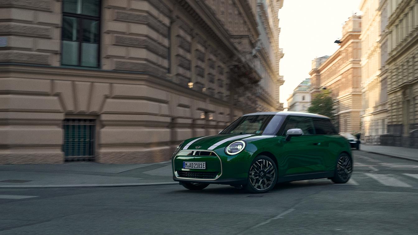 Ein vollelektrischer MINI Cooper Electric in klassischem British Racing Green mit weißem Dach und weißen Motorhaubenstreifen fährt dynamisch durch eine städtische Straße mit historischer Architektur.