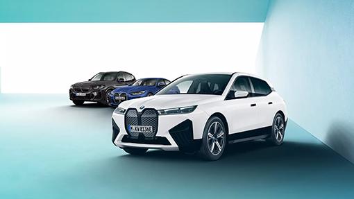 Weißer BMW iX mit blauem und dunkelgrauem BMW im Hintergrund.  Alle Fahrzeuge stehen in einem modernen, hellblauen Raum mit weißem Hintergrund.