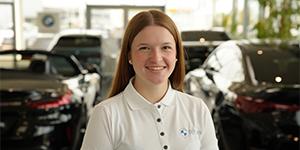 Kathlen Wehner, Auszubildene BMW Rhein.  Das Bild zeigt eine junge Frau in einem Autohaus, die direkt in die Kamera lächelt. Sie hat langes, glattes, braunes Haar, das offen über ihre Schultern fällt. Sie trägt ein weißes Poloshirt mit einem BMW-Logo auf der linken Brust.  Im Hintergrund sind verschwommen mehrere Autos zu erkennen. Eines der Autos ist ein schwarzes Cabrio auf der linken Seite des Bildes. Die Beleuchtung ist hell, was auf eine Innenaufnahme schließen lässt.