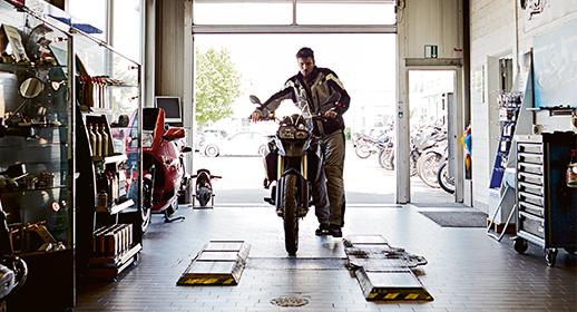 Dieses Bild ist aus dem Inneren einer Werkstatt in Richtung des hellen Einfahrtstors aufgenommen. Ein Mitarbeiter schiebt gerade eine große Reiseenduro (BMW GS) durch das Tor in die Werkstatt. Links sieht man Vitrinen mit Zubehör und Motoröl. Die Szene vermittelt den logistischen Aspekt des Kundenservice, besonders für die Wintermonate.