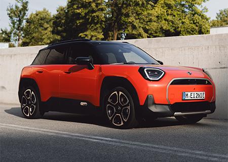 Lagerwagenaktion MINI BMW Rhein Ein leuchtend oranger Mini Aceman E, ein kompakter Elektro-SUV, steht auf einer Straße. Das Fahrzeug hat ein schwarzes Dach und schwarze Radkästen, die einen Kontrast zur orangen Karosserie bilden. Die Scheinwerfer sind eckig mit einer markanten Lichtsignatur. Im Hintergrund sind eine Betonwand und Bäume unter einem leicht bewölkten Himmel zu sehen.