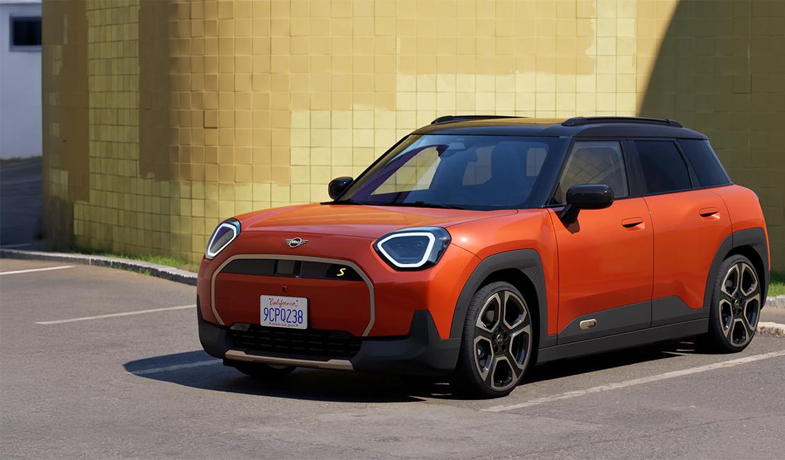 Das Bild zeigt den MINI Aceman Electric, den ersten rein elektrischen Crossover der Marke, in einer markanten orangefarbenen Lackierung. Im Gegensatz zum klassischen Cooper wirkt dieses Modell durch seine achteckigen Scheinwerfer, die robuste schwarze Radlaufverkleidung und die integrierte Dachreling deutlich kantiger und geländegängiger. Das Fahrzeug kombiniert ein schwarzes Kontrastdach mit aerodynamischen Felgen und ist vor einer strukturierten, olivgrünen Wand im hellen Sonnenlicht positioniert. Mit einer Länge von etwa 4,07 Metern schließt er die Lücke zwischen dem Cooper und dem Countryman und bietet Platz für fünf Personen sowie einen variablen Laderaum.