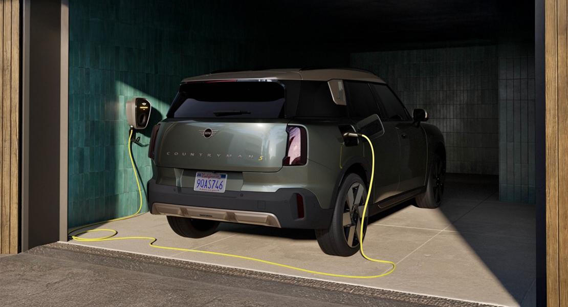 Das Bild zeigt einen modernen MINI Countryman S in einer stilvoll gestalteten Garage, während er gerade an einer Wandladestation aufgeladen wird. Das Fahrzeug ist in einem eleganten Olivgrün lackiert und präsentiert das markante, kantige Heckdesign der neuesten Modellgeneration, wobei der Modellname in großen Buchstaben und ein gelbes „S“ deutlich erkennbar sind. Ein leuchtend gelbes Ladekabel verbindet die Wallbox an der mit tiefblauen Kacheln verzierten Seitenwand mit dem Ladeanschluss des Wagens. Die Szene wird durch natürliches Licht von der Garagenöffnung her beleuchtet, was die metallische Oberfläche des Autos und das kalifornische Nummernschild hervorhebt und eine hochwertige, moderne Atmosphäre schafft.