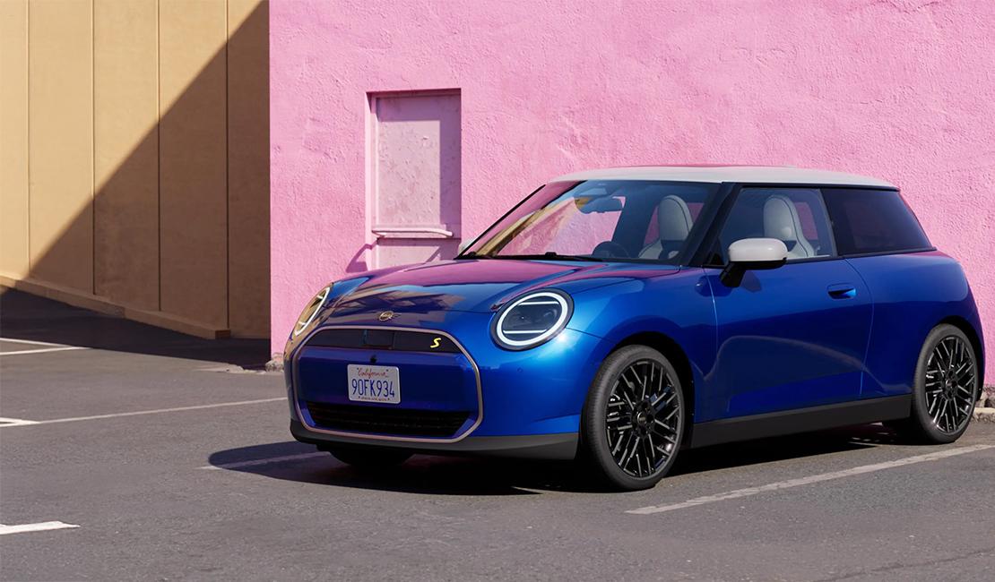  Das Bild zeigt einen modernen MINI Cooper Electric in einem kräftigen, glänzenden Blauton, der vor einer kontrastreichen Kulisse geparkt ist. Das Fahrzeug zeichnet sich durch sein charakteristisches Design mit runden LED-Scheinwerfern und einem weißen Dach sowie weißen Außenspiegeln aus. Besonders auffällig sind die filigranen, schwarzen Vielspeichen-Felgen.  Der Hintergrund besteht aus einer minimalistischen Architektur: Eine markante, leuchtend rosa Wand bildet einen starken farblichen Kontrast zum Blau des Autos, während ein angrenzender Wandabschnitt in dezentem Beige gehalten ist. Das harte Sonnenlicht erzeugt klare Schattenwürfe auf dem Asphalt und betont die glatten Oberflächen und Kurven des Fahrzeugs.