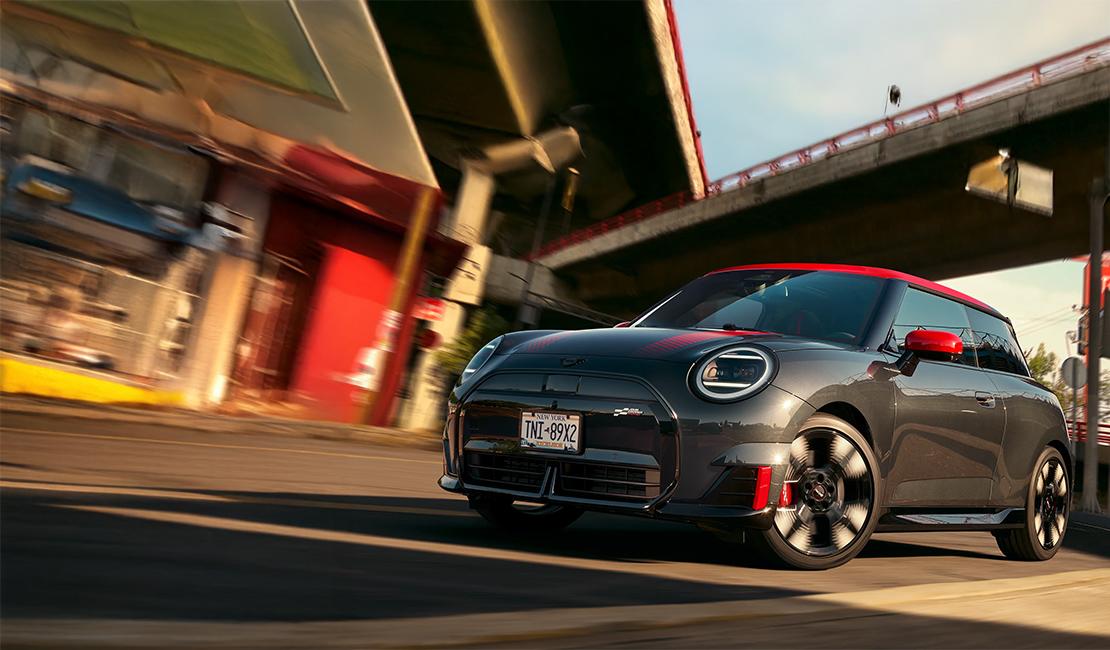  Das siebte Bild zeigt den MINI Cooper JCW Electric, die leistungsstarke Top-Variante des klassischen Dreitürers, in einer dynamischen Action-Aufnahme. Das Fahrzeug besticht durch seine sportliche Lackierung in dunklem Grau, kombiniert mit dem für die John Cooper Works Modelle charakteristischen roten Kontrastdach und roten Spiegelkappen. Spezifische JCW-Designmerkmale wie das exklusive Logo im Zielflaggen-Design, markante Aerodynamik-Anbauteile und große Leichtmetallräder mit roten Bremssätteln unterstreichen seinen Performance-Anspruch. In der gezeigten Fahrszene wird das typische „Gokart-Feeling“ visualisiert, das durch das sofortige Drehmoment des Elektroantriebs und ein besonders sportlich abgestimmtes Fahrwerk erreicht wird.
