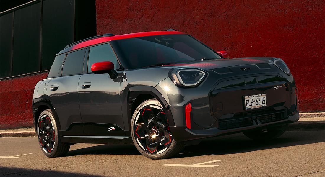 Ein glänzender, dunkelgrauer MINI John Cooper Works Aceman SUV mit rotem Dach und roten Außenspiegeln steht auf einem Bürgersteig vor einer roten Wand. Das Fahrzeug trägt das New Yorker Kennzeichen "GLH-62J5" und weist sportliche rote Akzente an der Front und an den Felgen auf.