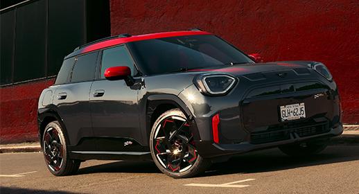 Ein glänzender, dunkelgrauer MINI John Cooper Works Aceman SUV mit rotem Dach und roten Außenspiegeln steht auf einem Bürgersteig vor einer roten Wand. Das Fahrzeug trägt das New Yorker Kennzeichen "GLH-62J5" und weist sportliche rote Akzente an der Front und an den Felgen auf.