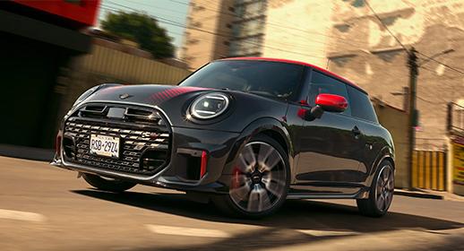 Ein glänzender, dunkelgrauer MINI John Cooper Works mit rotem Dach, roten Spiegelkappen und roten Akzenten an der Front, fährt dynamisch auf einer Straße mit unscharfem Hintergrund, der die Bewegung betont. Das Fahrzeug trägt ein New Yorker Kennzeichen "RSB-2924" und hat eine sportliche Optik mit roten Zierstreifen auf der Motorhaube.