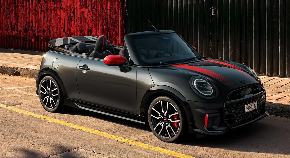 Ein glänzendes, dunkelgraues MINI John Cooper Works Cabriolet mit geöffnetem schwarzen Stoffverdeck, roten Spiegelkappen und roten Rennstreifen auf der Motorhaube. Das Fahrzeug steht auf einem Bürgersteig vor einer roten Wand und hat sportliche schwarze Felgen mit roten Bremssätteln.