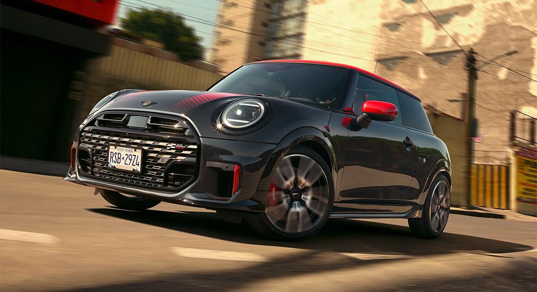 Ein glänzender, dunkelgrauer MINI John Cooper Works mit rotem Dach, roten Spiegelkappen und roten Akzenten an der Front, fährt dynamisch auf einer Straße mit unscharfem Hintergrund, der die Bewegung betont. Das Fahrzeug trägt ein New Yorker Kennzeichen "RSB-2924" und hat eine sportliche Optik mit roten Zierstreifen auf der Motorhaube.