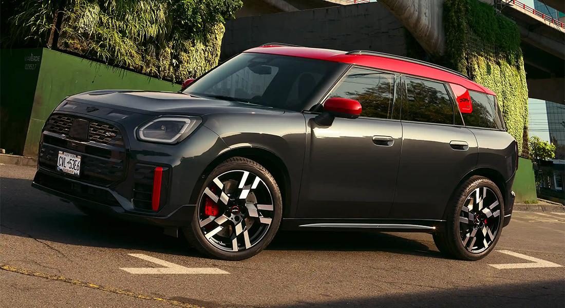 Ein dunkelgrauer MINI JCW Countryman All4 mit rotem Dach und roten Spiegelkappen steht auf einer Straße. Das Auto ist schräg von vorne zu sehen, mit Blick auf die Fahrerseite. Im Hintergrund sind grüne Pflanzen und eine Gebäudestruktur erkennbar.