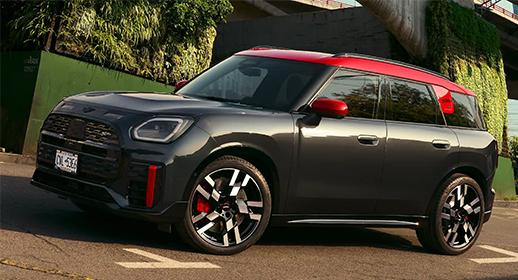 Ein dunkelgrauer MINI JCW Countryman All4 mit rotem Dach und roten Spiegelkappen steht auf einer Straße. Das Auto ist schräg von vorne zu sehen, mit Blick auf die Fahrerseite. Im Hintergrund sind grüne Pflanzen und eine Gebäudestruktur erkennbar.