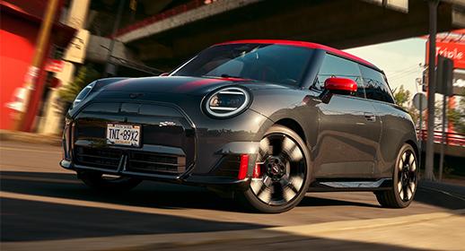 Ein glänzender, dunkelgrauer MINI John Cooper Works Electric mit rotem Dach, roten Spiegelkappen und roten Akzenten an der Front. Das Auto ist in Bewegung auf einer Straße bei sonnigem Wetter, mit verschwommenem Hintergrund, der urbane Elemente andeutet.