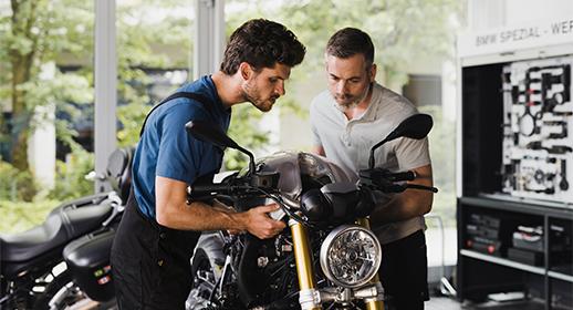 In dieser Aufnahme begutachten zwei Personen – ein jüngerer Mechaniker und ein älterer Kollege oder Kunde – sehr genau die Frontpartie eines Motorrads. Sie wirken fokussiert und besprechen technische Details. Dies unterstreicht die technische Expertise und die gründliche Diagnose, die nach Stürzen oder Unfällen notwendig ist.