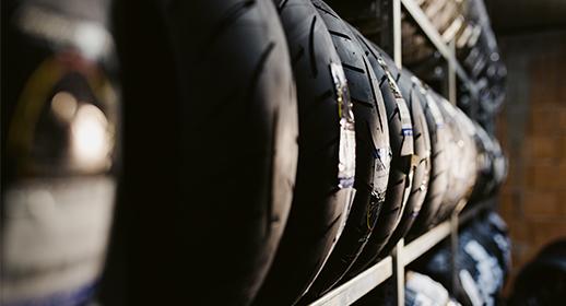 Das Bild zeigt eine Nahaufnahme von fabrikneuen Motorradreifen, die in einem Lagerregal aufreiht sind. Die Tiefenschärfe ist so gewählt, dass die Profilstruktur der vorderen Reifen deutlich erkennbar ist. Es symbolisiert die Verfügbarkeit von Verschleißteilen und die Sicherheit durch frisches Material.