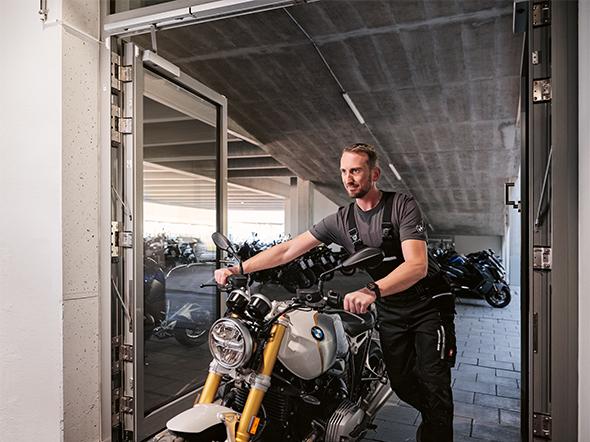 Ein Mitarbeiter schiebt eine moderne BMW R nineT durch ein Werkstattportal; im Hintergrund ist ein gut gefüllter Parkplatz mit verschiedenen Modellen (u.a. Scooter) zu sehen.