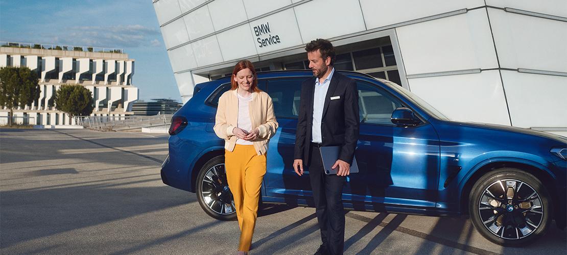 BMW Service Inclusive Ein Mann im Anzug mit einem Tablet und eine Frau in gelber Hose und beigefarbener Jacke mit einem Smartphone gehen lächelnd auf dem Parkplatz vor einem modernen BMW Servicegebäude. Ein blauer BMW SUV ist im Hintergrund geparkt.