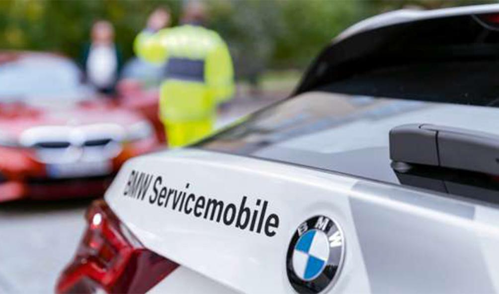 Das Bild zeigt eine Nahaufnahme des Hocks bzw. der Seite eines weißen BMW Servicemobils, während im unscharfen Hintergrund ein Mitarbeiter in gelber Warnkleidung sowie ein rotes Auto zu erkennen sind.