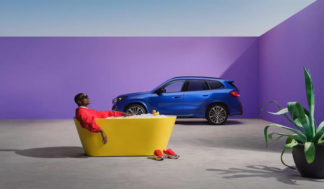 Blauer BMW X1 in Seitenansicht vor einer violetten Wand. Im Vordergrund entspannt sich ein Mann in einer gelben Badewanne.