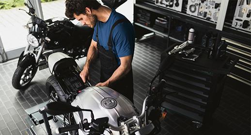 Das Bild zeigt einen Mechaniker in blauer Arbeitskleidung, der konzentriert an einem silbernen Motorrad arbeitet. Er scheint gerade das Heck oder die Sitzbankpartie zu demontieren oder zu prüfen. Im Hintergrund ist eine saubere, gut organisierte Werkstatt mit Werkzeugwagen und einem weiteren schwarzen Motorrad zu sehen. Der Fokus liegt hier auf der Präzision und Detailarbeit bei Reparaturen.